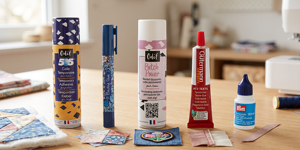 Les cinq familles de colles textiles côte à côte : spray colle temporaire, stylo colle, colle thermocollante en spray et ruban, tube de colle textile permanente, flacon de colle anti-effilochage