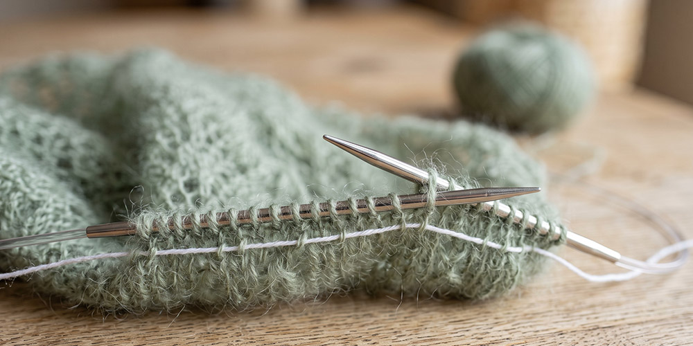 Fil de sécurité blanc passé dans les mailles d'un tricot en mohair vert sauge sur aiguilles circulaires en métal